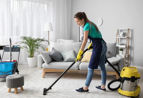A friendly, professional house cleaner in Toronto vacuuming a light-colored living room rug, demonstrating residential cleaning services in a modern home environment.