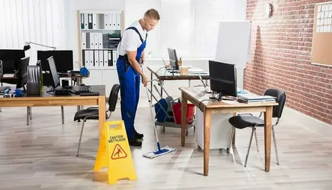 A professional male cleaner wearing blue uniform and a 'Caution Wet Floor' sign mopping a wooden floor near office desks, highlighting safe and thorough commercial cleaning services in Toronto by Eco Techno Clean.