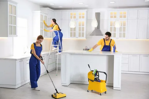 Three Eco Techno Clean staff performing detailed kitchen house cleaning in Toronto, including one person on a ladder cleaning cabinets and others wiping down counters and mopping the floor.
