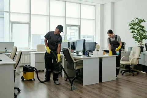 Two professional cleaners performing office cleaning in a bright Toronto open-concept office, with one vacuuming the floor and the other wiping down a desk.