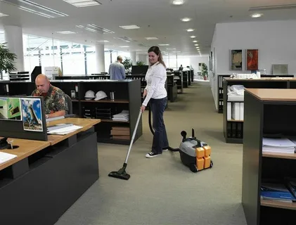 Female cleaner vacuuming the carpeted floor next to an employee working at his desk, demonstrating flexible office cleaning during business hours in a large Toronto office.