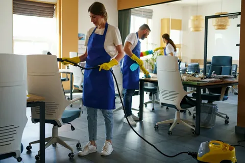A team of three 'Eco Techno Clean' staff members in blue aprons and yellow gloves using a steam cleaner on an office chair and wiping down surfaces in a commercial office.